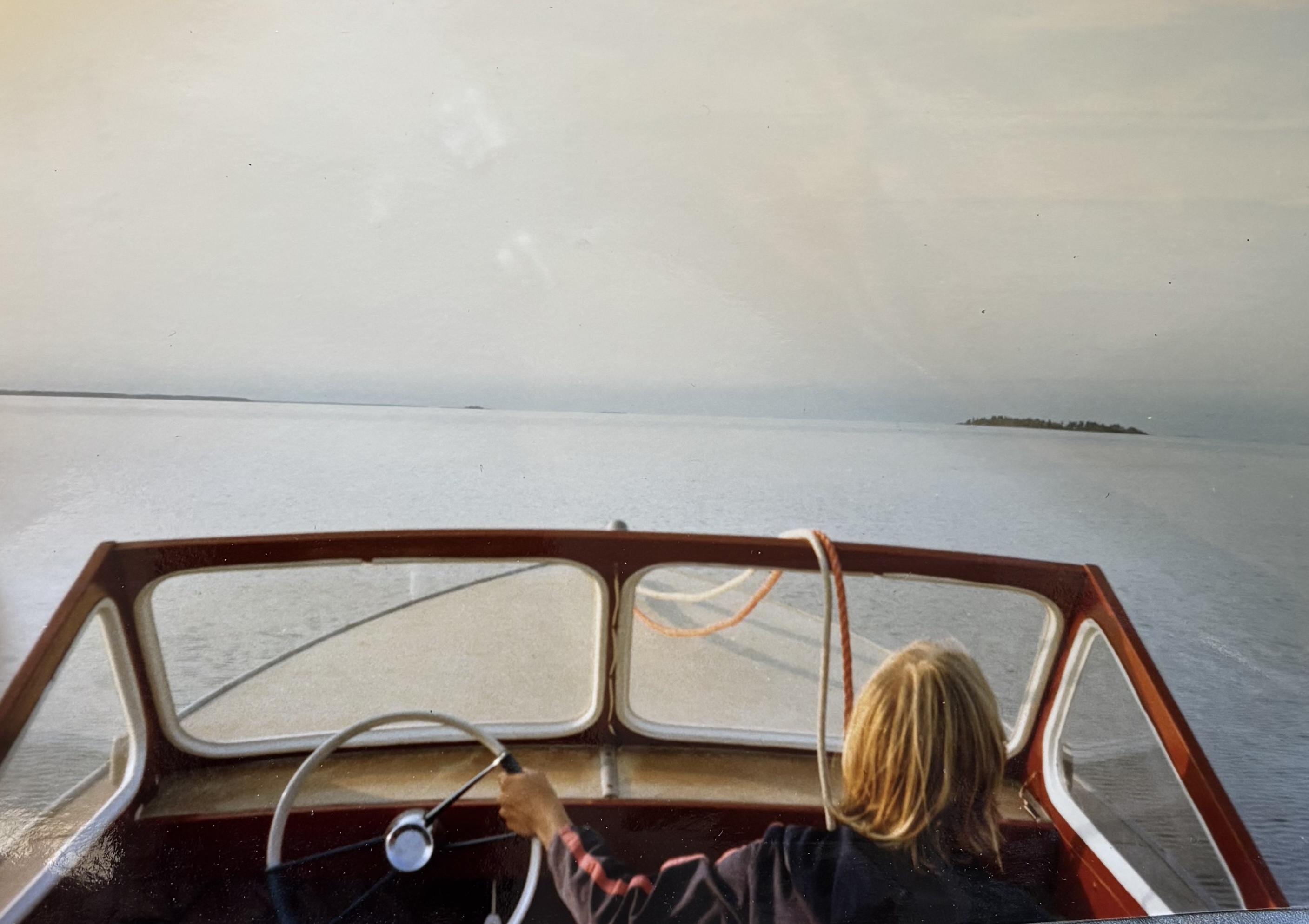 A person with blonde hair pilots a speedboat with one hand, the ocean in the background.